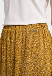 Gonna floreale in giallo senape con fiori bianchi e viola, design plissettato, vita elastica e una piccola etichetta metallica.