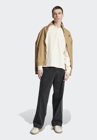 Chaqueta ligera beige, polo de manga larga crema, vaqueros de pierna ancha negros, zapatillas blancas; el modelo está de pie en un ambiente neutro.