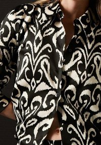 Blusa bianca e nera con un intricato motivo a spirale, realizzata in tessuto morbido, con colletto classico e chiusura a bottoni.