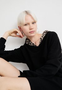 Čierna mikina s leopardím vzorom na kapucni, so strihom do V a dlhými rukávmi. Hladká textúra a voľný strih.