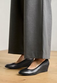 Chaussures compensées en cuir noir avec un bout arrondi et une texture lisse, associées à un pantalon large gris, posées sur un sol en bois.