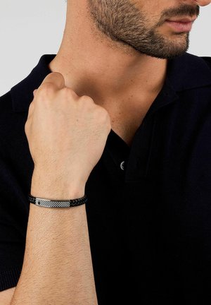 Uomo che indossa un braccialetto nero intrecciato in pelle con una piastra d'argento testurizzata, vestito con una polo nera, pugno alzato vicino al mento.