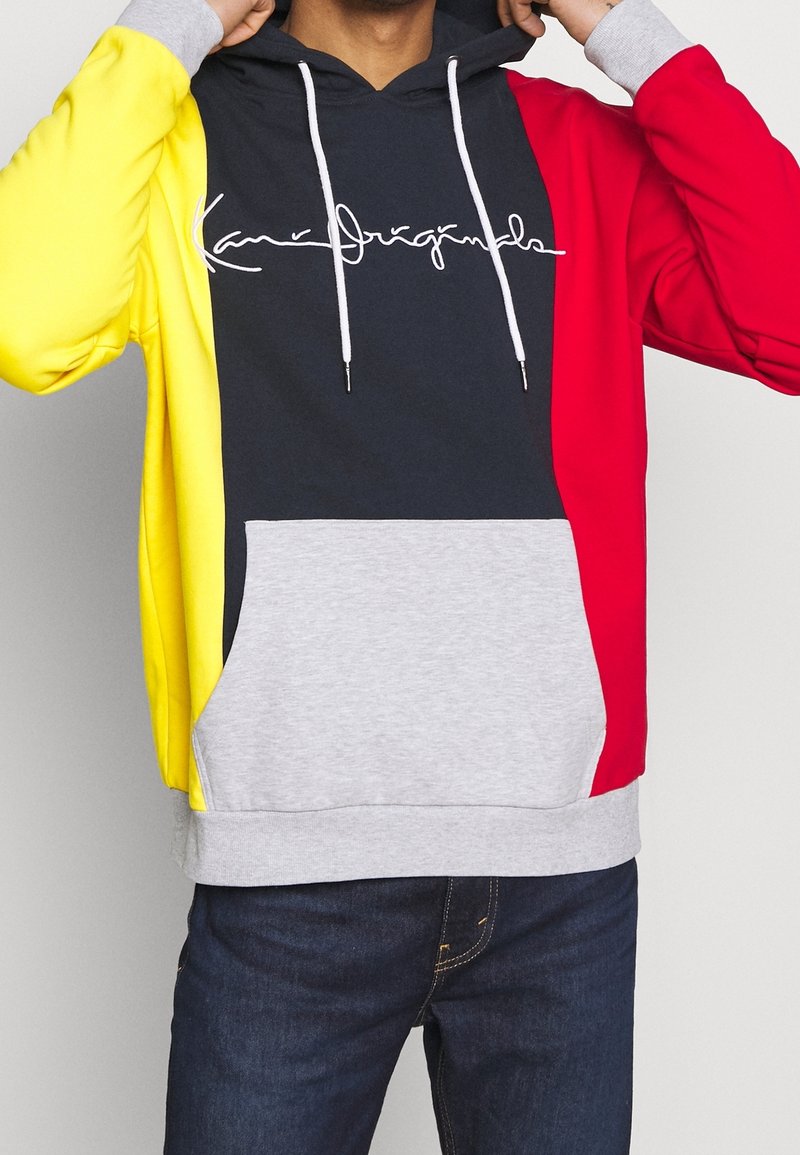 Mikina s kapucí s tmavě modrým středovým panelem, žlutým levým rukávem, červeným pravým rukávem a šedou kapsou. Obsahuje bílé šňůrky na stažení a vytištěné logo.