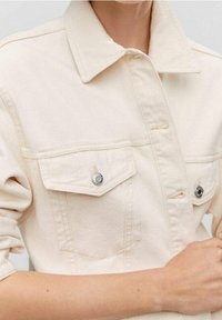 Veste en denim beige clair avec un col classique, deux poches avant, boutons en métal argenté et manches retroussées. Texture lisse dans l'ensemble.