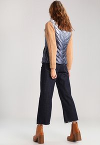 Vadderad väst med en glänsande, mörkblå framdel och stickade beige ärmar, i kombination med mörka vida jeans och bruna klackiga ankelboots.