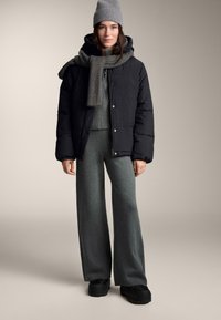 Čierna puff jacket, šedý pletený sveter, dlhé sivé nohavice a šedá čiapka. Oblečenie obsahuje rebrovaný šál a čierne topánky.