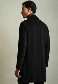 Sort frakke med en glat tekstur, skræddersyet pasform og en klassisk notch-krave, præsenteret på en model set fra ryggen mod en neutral baggrund.