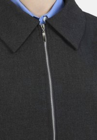 Giacca senza colletto di colore grigio scuro con una chiusura frontale metallica, realizzata in tessuto testurizzato, con una camicia azzurra chiara visibile sotto.