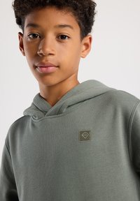 Olivegrüner Kapuzenpullover mit weicher Textur, ausgestattet mit einem kleinen quadratischen Logo auf der Brust und einem lässigen Schnitt. Dezente Stickdetails sind sichtbar.