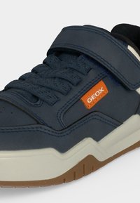 Gros plan sur une basket bleu marine avec des lacets, une sangle velcro, une semelle blanche, une semelle extérieure marron et une étiquette orange de la marque Geox sur le côté.