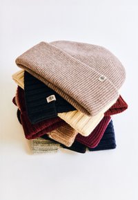 Stack of knit beanies in various colors: beige, cream, black, burgundy, gray, and navy, featuring ribbed texture and logo tag on each.