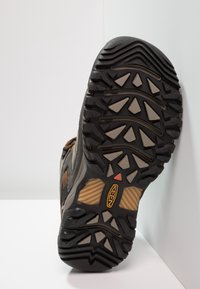 Schwarz-grüner Wander-Schuhsohle mit einer strukturierten Gumm OBERFLÄCHE und geometrischen Profilmustern. Mit einem gelben Akzent und einem roten Indikator.