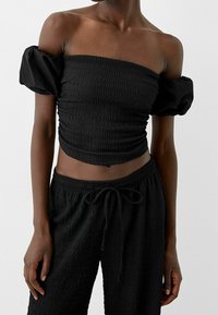 Haut noir épaules dénudées avec texture smockée et manches ballon, assorti d'un pantalon noir ample avec une taille à cordon.