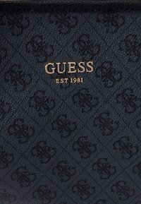 Tissu noir texturé avec un motif floral foncé répété et un texte doré centré indiquant "GUESS EST 1981."