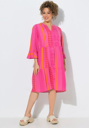 Kobieta o kręconych włosach, ubrana w jasno-różową sukienkę do kolan z geometrycznymi pomarańczowymi i czerwonymi wzorami, w połączeniu z beżowymi sandałami na platformie.