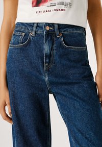 Temno modre kavbojke iz jeansa z visokim pasom, petimi žepi, kontrastnim šivom in gumbom s kovinsko medeno obdelavo.