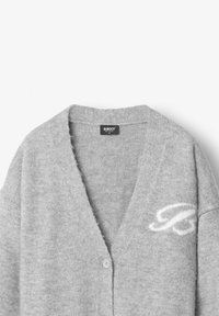 Šedý svetr s výstřihem do V, měkkou texturou, zapínáním na knoflíky a bílým logem na levé straně. Vyroben z příjemného materiálu.