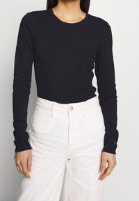 Musta pitkähihainen paita, jossa pyöreä pääntie, istuva malli ja pehmeä materiaali, yhdistettynä valkoisiin korkeavyötäröisiin housuihin, joissa on nappisuljenta.