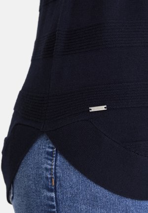 Dunkelblauem Strickpullover mit strukturierten horizontalen Streifen und einem kleinen rechteckigen Metall-Logoschild über blauen Jeans.