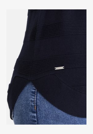 Dunkelblauem Strickpullover mit strukturierten horizontalen Streifen und einem kleinen rechteckigen Metall-Logoschild über blauen Jeans.