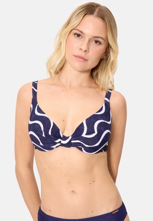 Mujer rubia con top de bikini azul marino con rayas onduladas blancas y parte inferior de bikini azul marino sólido a juego sobre fondo claro.