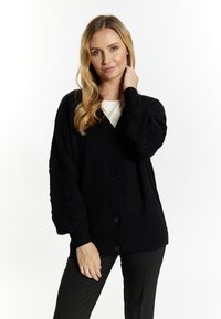 Cardigan nero a maglia con bottone frontale, caratterizzato da maniche a palloncino testurizzate e una vestibilità rilassata, indossato sopra un top chiaro.