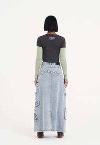 Lange Jeansrock mit Cargo-Taschen, hellblauer Waschung und gesticktem Design. Kombiniert mit einem grau-grünen Lagenoberteil, das einen Text-Print enthält.