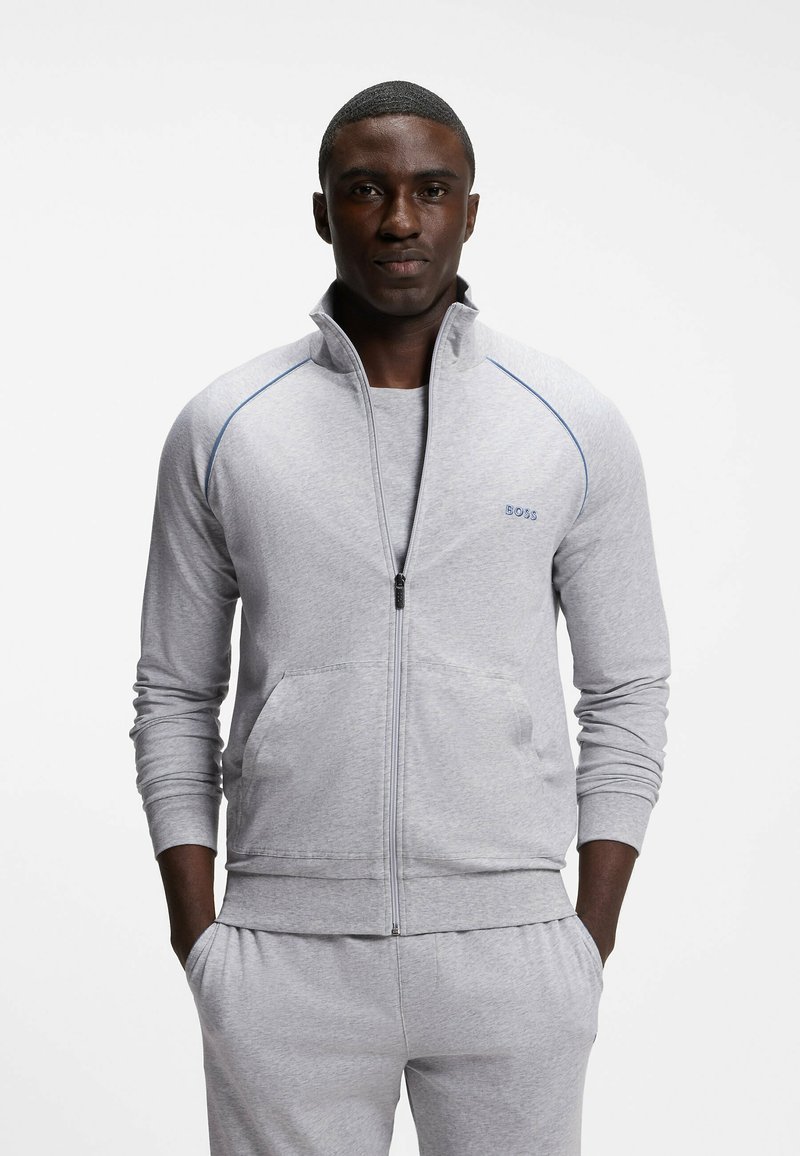 Homme portant une veste zippée gris clair et un pantalon assorti, se tenant les mains dans les poches devant un fond blanc uni.