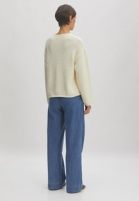 Cremefarbener, strukturierter Strickpullover mit lockerer Passform, kombiniert mit weiten blauen Denim-Hosen, von hinten vor einem hellen Hintergrund präsentiert.
