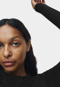 Czarny top z długim rękawem o dopasowanym kroju, gładkiej fakturze i okrągłym dekolcie, noszony przez osobę z falującymi ciemnymi włosami, podkreślający twarz.