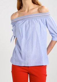 Helles Blau, schulterfreies Blusenshirt mit strukturiertem Smocking am Kragen, kurzen Ärmeln und Bindebändern. Getragen mit roten Hosen.