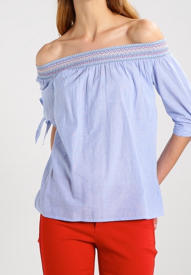 Helles Blau, schulterfreies Blusenshirt mit strukturiertem Smocking am Kragen, kurzen Ärmeln und Bindebändern. Getragen mit roten Hosen.