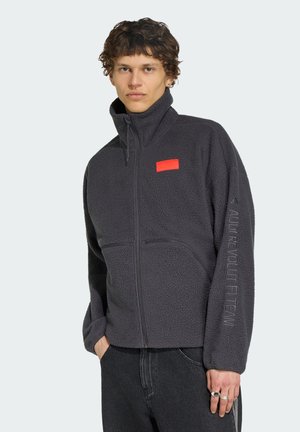 Junger Mann trägt eine dunkelgraue Fleecejacke mit durchgehendem Reißverschluss, einem roten Aufnäher auf der Brust und Text am linken Ärmel, steht mit teilweise in den Taschen steckenden Händen.