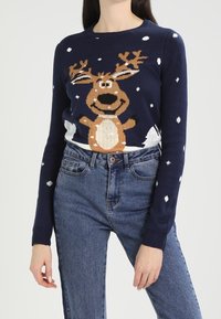 Granatowy sweter dziany z motywem renifera z porożem, brązowym tułowiem, kremowym brzuchem oraz białymi akcentami w kształcie płatków śniegu. Lekka struktura tkaniny.