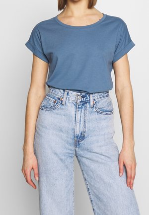 Kvinde iført en blå t-shirt med korte ærmer og opkrogede manchetter, stoppet ned i lyseblå, højtaljede denimbukser mod en ensfarvet baggrund.