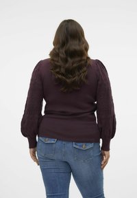 Donna con lunghi capelli castani mossi, che indossa un top lungo a maniche lunghe di colore viola con texture e jeans blu, in piedi di spalle contro uno sfondo neutro.