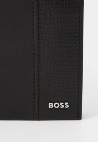 Pannello in pelle nera testurizzata con bordi cuciti e piccolo logo "BOSS" argentato nell'angolo in basso a destra su superficie bianca.