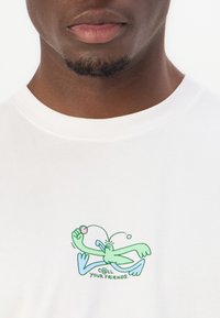 T-shirt blanc avec un personnage de dessin animé vert tenant un ballon, sous-titré "c@ll YOUR FRIENDS." Le design est centré sur la poitrine.