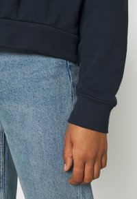 Una mano adulta appoggiata accanto a jeans in denim blu e a una felpa a maniche lunghe blu navy con maniche a cuffia su uno sfondo chiaro.