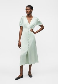 Ženska nosi svetlo zeleno midi obleko s kratkimi rokavi in gumbi spredaj ter črne balerinke, stoji z roko na boku.