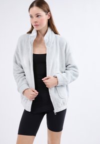 Veste en polaire gris clair avec un col montant, des manches longues et des poches latérales. Texture lisse, coupe décontractée et design à l'avant ouvert.