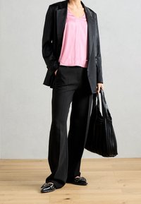 Woman wearing a black blazer, pink blouse, black wide-leg trousers, black studded loafers, and carrying a large pleated black handbag.