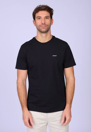 Homme debout face à l'avant, portant un t-shirt noir uni avec un petit logo animal blanc sur la poitrine et un pantalon de couleur claire.