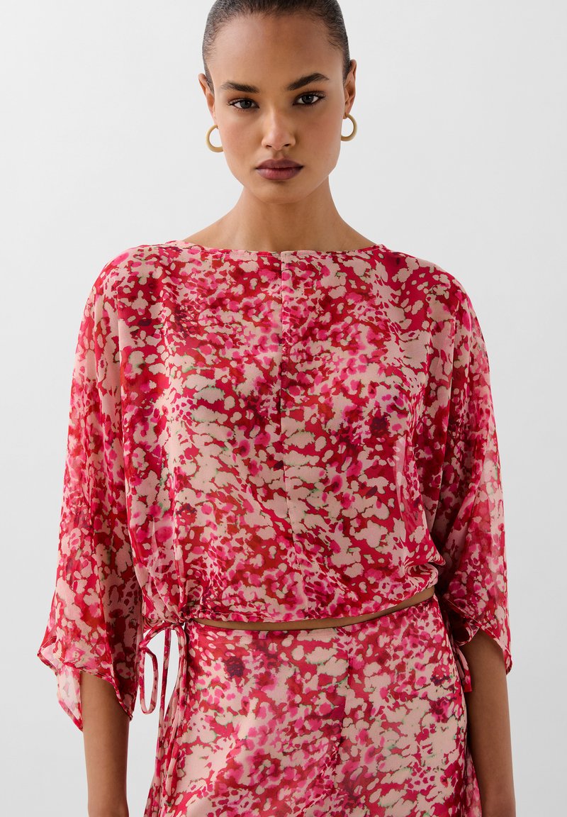 Blouse florale en tissu transparent, avec un mélange de couleurs rose, rouge et beige. Coupe ample avec des manches chauve-souris et un nœud à la taille.