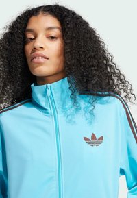 Hellblaues Zip-Up-Jackett mit Stehkragen, das braune Streifen an den Schultern und ein braunes Adidas-Logo auf der Brust aufweist. Glatte Textur.
