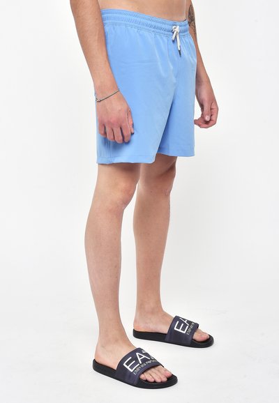 Pantaloni da bagno azzurri chiari con vita elastica e cordoncino. Abbinati a sandali neri con fascia logo bianco.