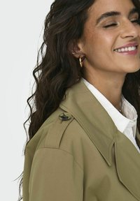 Olivgrön trenchcoat med en strukturerad krage och knappdetalj, bärs över en vit skjorta. Långt, vågigt mörkt hår delvis synligt.