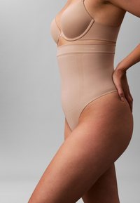 Kvinde iført nude høj talje formgivende undertøj og matchende bh, stående sidelæns med den ene hånd på hoften mod en ensfarvet baggrund.
