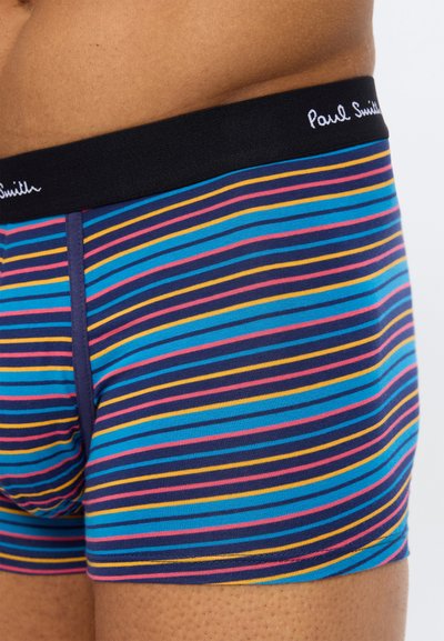 Κοντινό πλάνο ενός ατόμου που φοράει ριγωτά σλιπ μποξέρ σε μπλε, ροζ και κίτρινο χρώμα με μαύρη ζώνη μέσης που φέρει το λευκό κείμενο "Paul Smith".