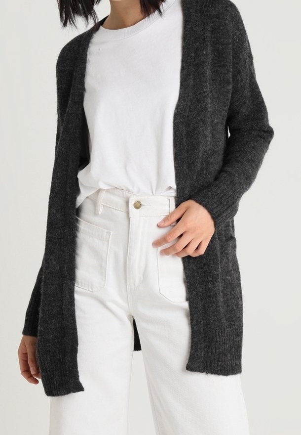 Cardigan en maille gris foncé avec un devant ouvert, des manches longues et une finition texturée, porté par-dessus un t-shirt blanc et un pantalon de couleur claire.